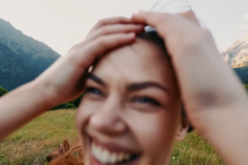 Young woman with long hair smiles joyfully while holding her head outdoors in a mountainous landscape during a bright day.