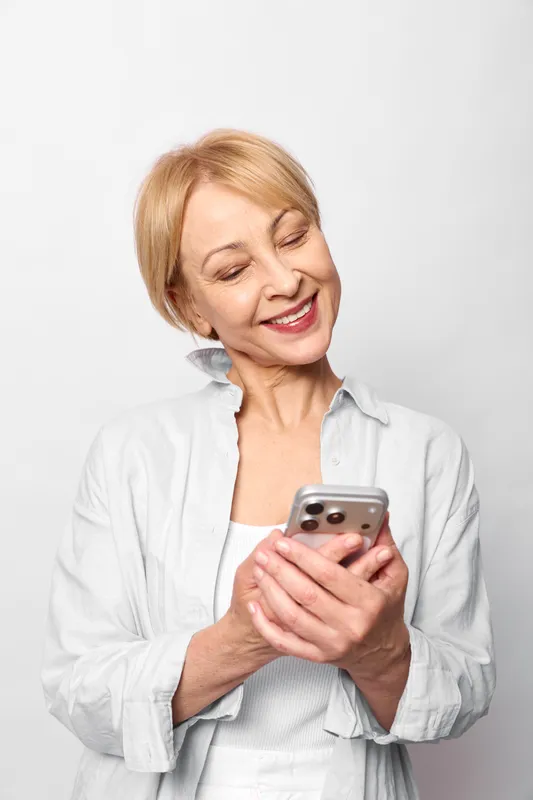 Elderly woman smiling while using a smartphone indoors, close view on a white background. Communication technology concept, happy mood, senior lifestyle.