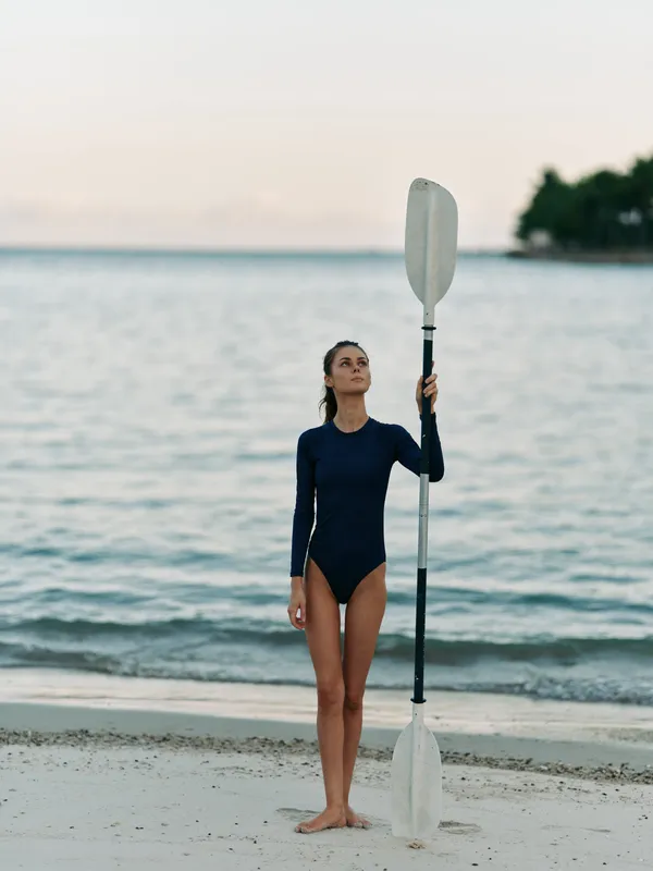 A woman in a dark navy swimsuit stands barefoot on a sandy beach, holding a tall paddle upright as calm waves and a serene horizon loom behind.