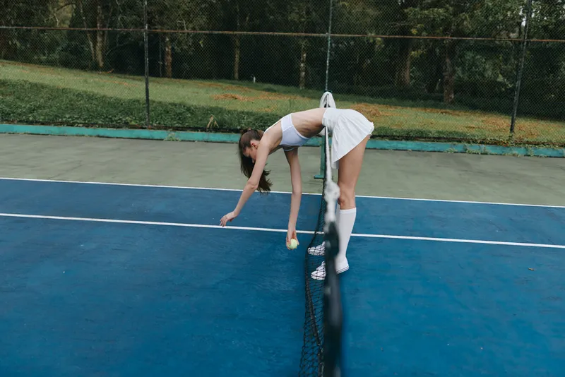 Young female tennis player in sporty white attire picking up a tennis ball on a green court surrounded by nature, embodying focus and determination