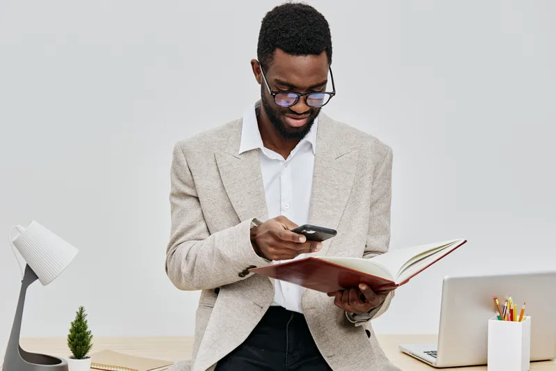 Professional young man using smartphone and reading a notebook while standing at a desk with a laptop and supplies, in a modern office environment. Business communication and technology concept, full 