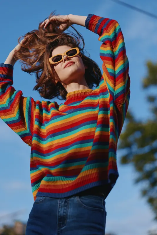 Woman in her mid 20s wearing a rainbow sweater and yellow sunglasses outdoors, smiling and flipping hair, casual outfit with jeans, confident playful pose for fashion and lifestyle.