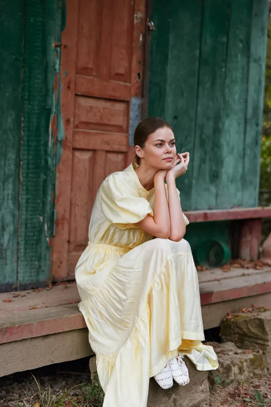 Woman in a light yellow dress sits on outdoor steps beside weathered doors, gazing into the distance with a thoughtful, calm expression.