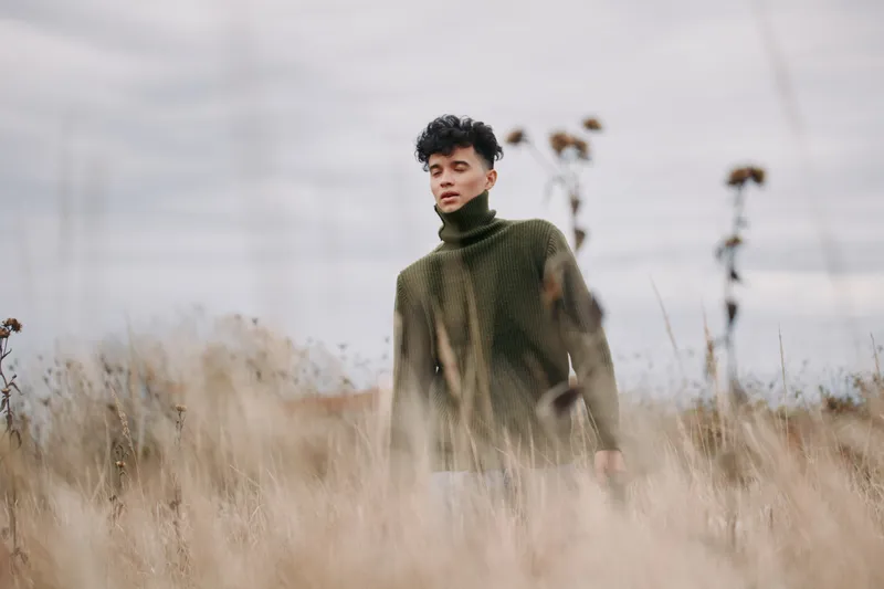 A young man in a knitted turtleneck walks through tall dry grass, a candid portrait emphasizing natural authenticity and credible, unposed presence beneath a muted, overcast sky.