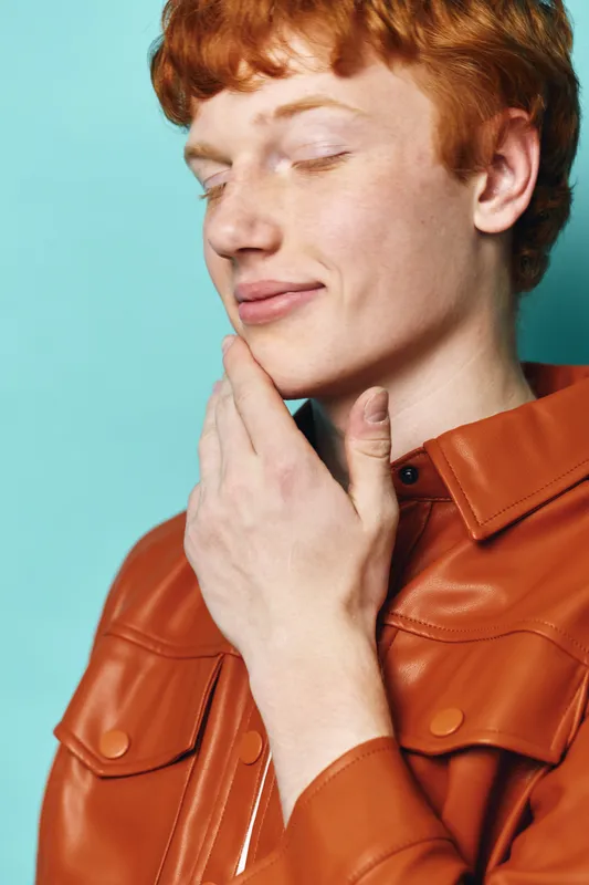 Young man with red hair wearing brown leather jacket posing with eyes closed and hand touching chin on blue background. Fashion, portrait and thoughtful mood of male model.