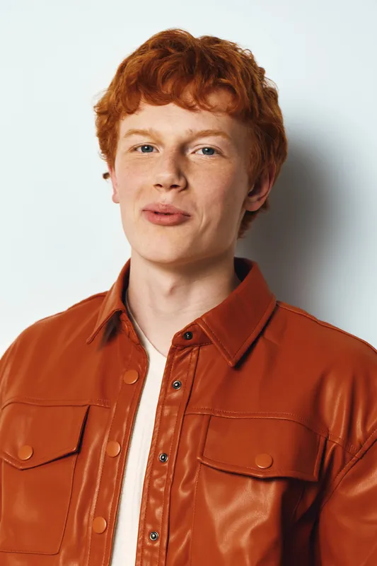 Young man with red hair wearing orange leather jacket and white shirt standing against white background looking at camera with slight smile and neutral expression.