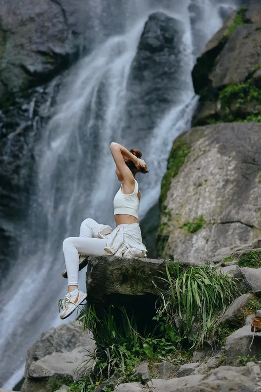 Woman practicing yoga poses near a waterfall wearing white sportswear, surrounded by rocks and lush green plants in a serene natural setting.