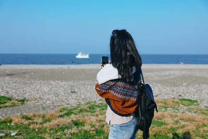 Woman on a beach with a backpack and camera captures photography of the sea, coastline, and shore, presenting ocean views and a wide landscape in a calm outdoor moment