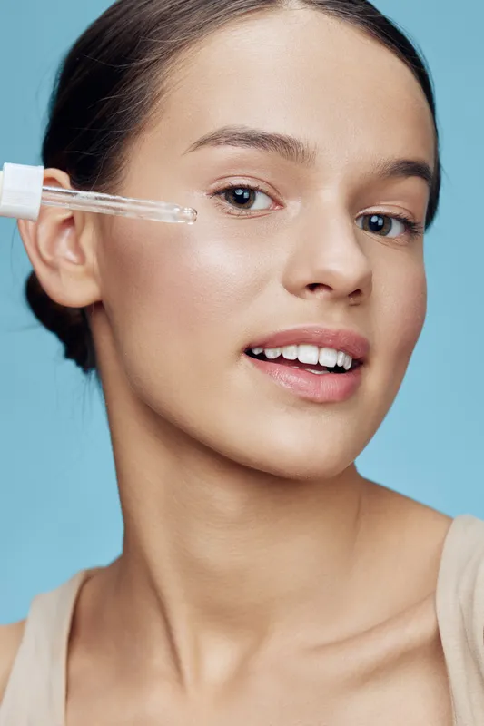 Young woman applying skincare serum, showcasing clear skin and a natural glow against a soft blue background, representing beauty and self care