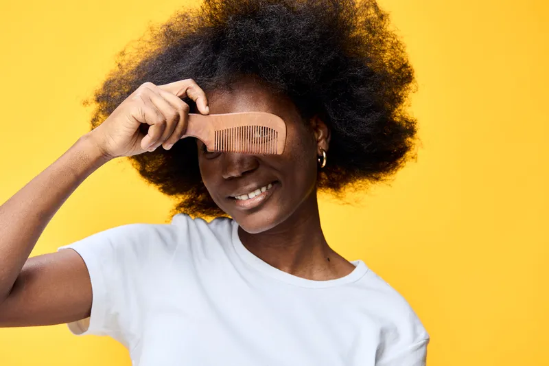 A joyful African model playfully using a comb to hide her eyes, showcasing natural hair against a vibrant yellow background, perfect for promoting hairstyling and self expression