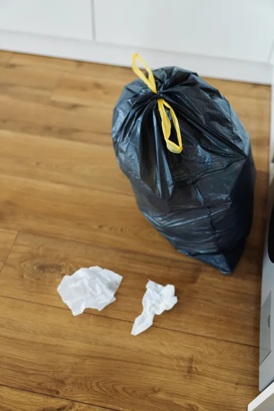 Trash garbage waste bag black rubbish on wooden floor near kitchen, tied plastic sack with yellow tie and crumpled tissues, domestic household disposal and cleaning scene.