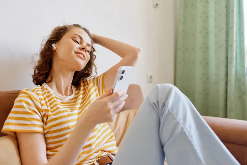 Brightly lit living room scene shows a young woman relaxing on a cozy sofa at home, casually wearing a striped tee and light jeans, scrolling her phone with a content smile.