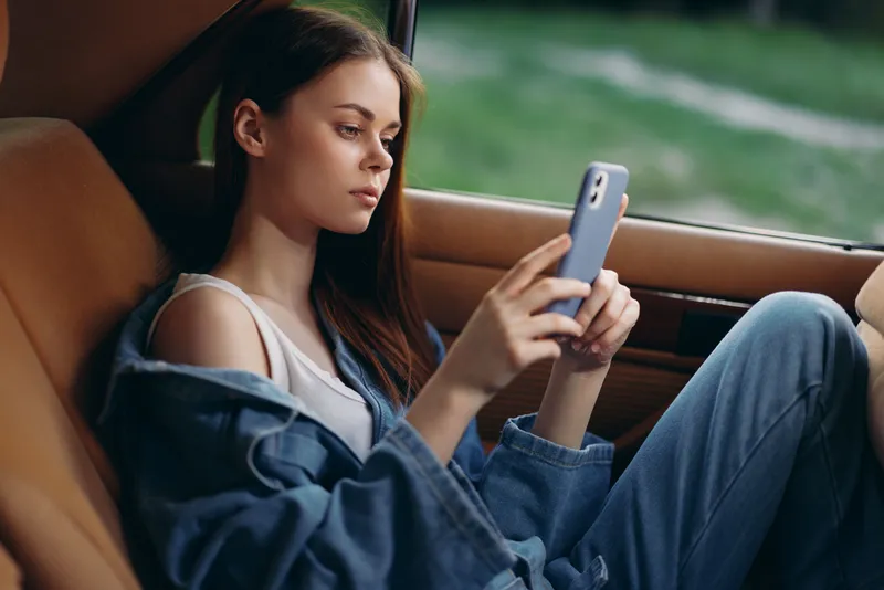 Young woman in casual denim outfit using smartphone in car, showcasing modern lifestyle and urban themes.