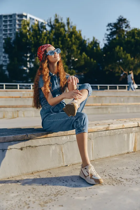 Lifestyle woman in modern boho-western denim street style wearing sunglasses and bandana, enjoying sunny outdoor urban setting with film color mood.