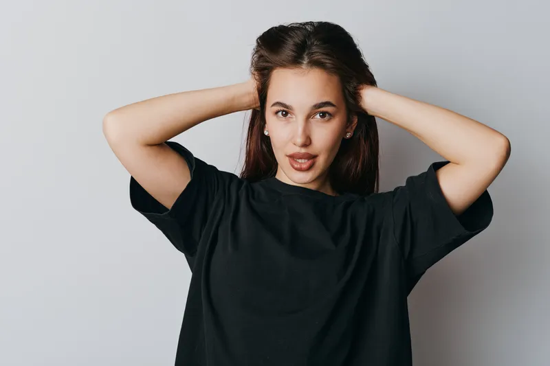 Young woman in black t shirt posing with hands behind head looking confident and casual against gray background in studio portrait with natural makeup and relaxed expression.