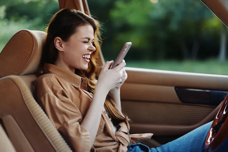 Happy young woman in a car enjoying texting on her smartphone during a sunny day, casual outfit and relaxed mood, vibrant greenery in the background.