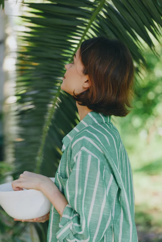 A woman outdoors wearing a striped green shirt holds a white bowl, with soft sunlight filtering through palm leaves.