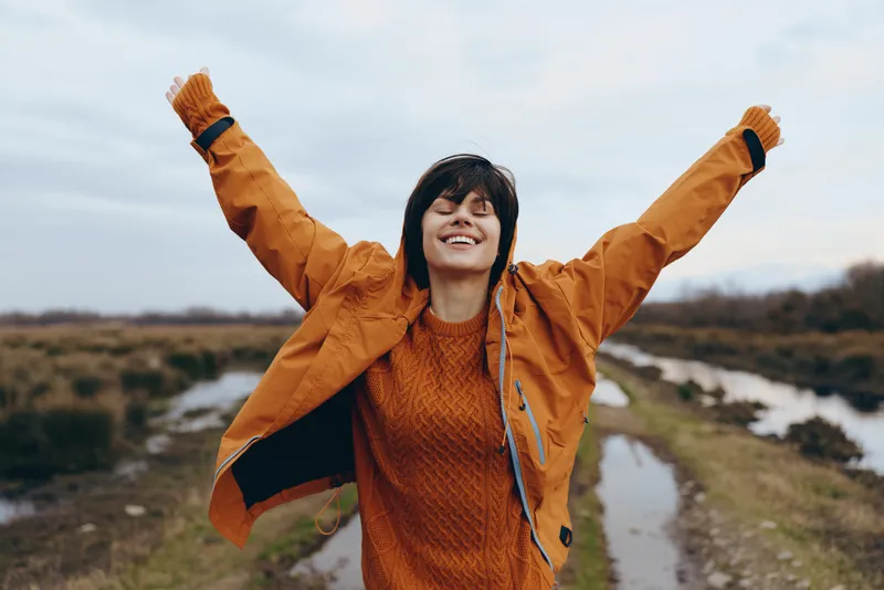 Woman lifestyle outdoors, victory pose with raised arms, orange jacket and knit sweater, smiling face toward sky, walking on riverside path in nature, fresh air mood, autumn landscape