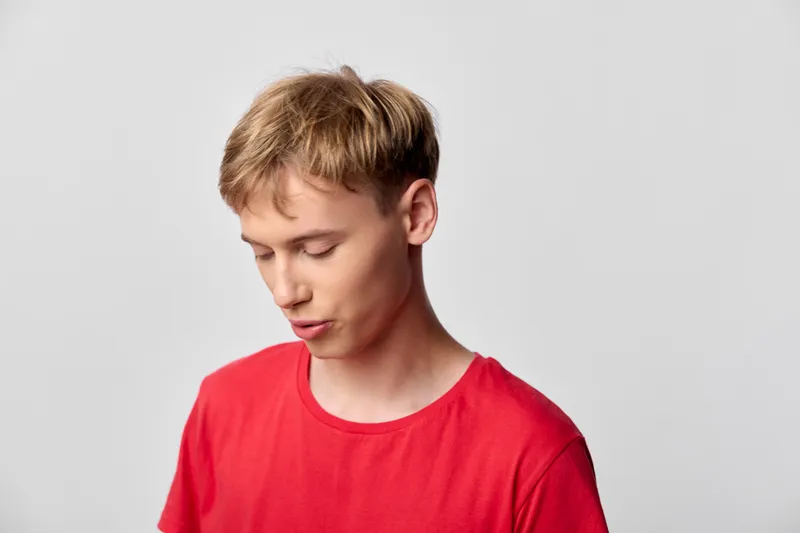 Young man in a red shirt with a thoughtful, downcast mood, casual pose in a neutral studio setting, close up composition, simple fashion and calm expression