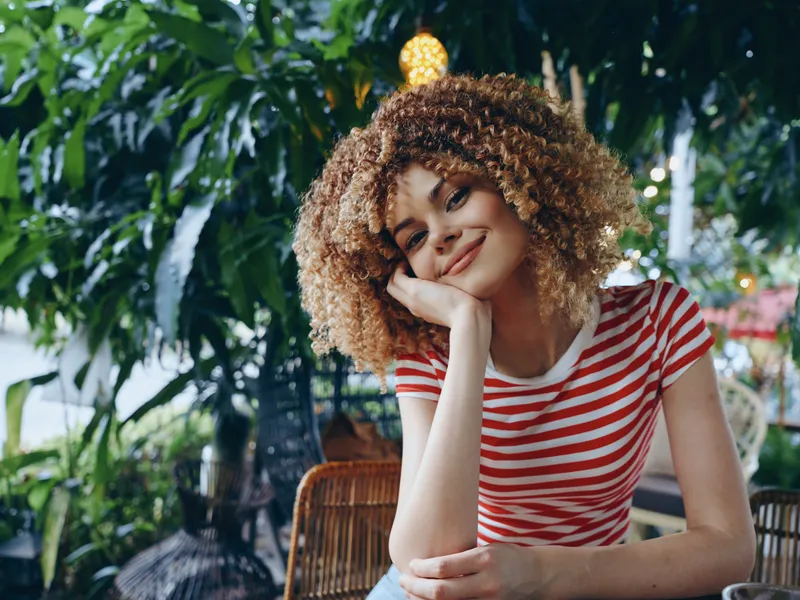 Portrait of a cheerful young woman with curly hair and striped shirt, resting her chin on her hand in a sunlit cafe setting, against a lush background of plants and soft lighting.