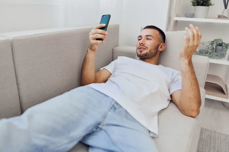 Relaxed man using smartphone while lounging on a cozy sofa, showcasing modern lifestyle and digital engagement. Ideal for concepts around leisure and technology.