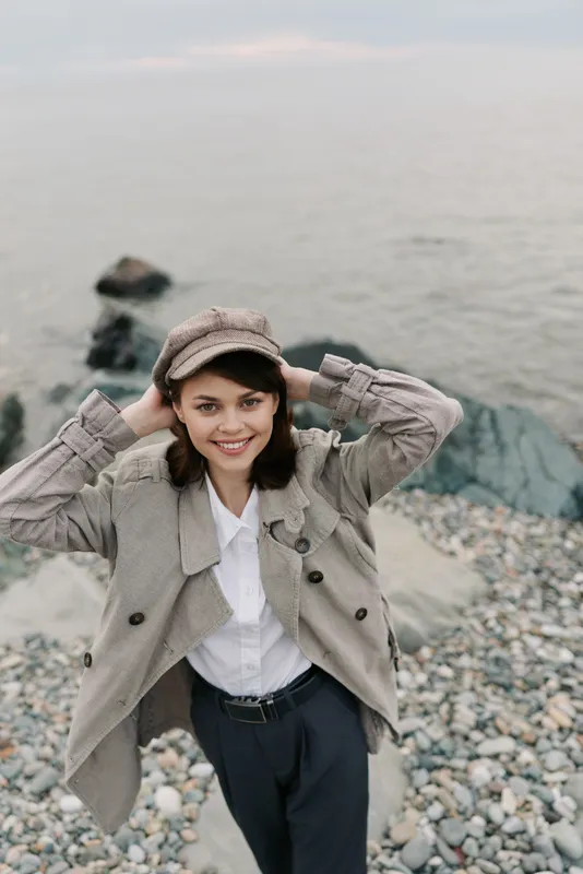 Young woman in a beige coat and cap smiles while standing on a rocky beach near calm water. Casual autumn outfit and natural outdoor lifestyle concept.