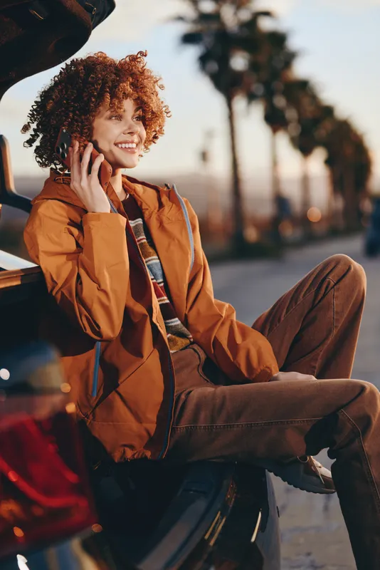 Woman lifestyle outdoors, smiling holding a phone near a car, curly hair visible, wearing an orange jacket in golden sunset light, relaxed street pose, soft, warm bokeh background