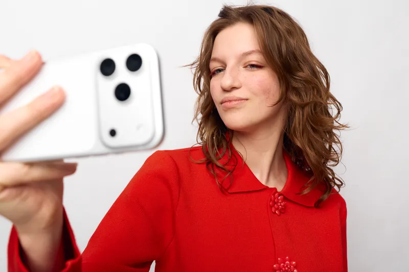 Young woman with wavy brown hair wearing red sweater takes selfie with white smartphone on white background. Concept of social media, technology, and casual lifestyle portrait.