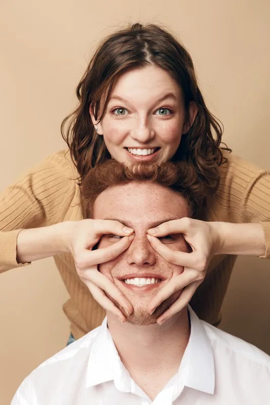 Happy young woman playfully covers smiling man face with hands in heart shape. Casual couple in beige and white clothes on neutral background. Concept of love, fun, and relationship.