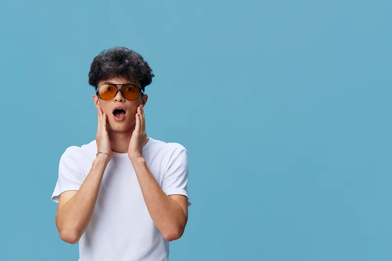 surprised man in white t-shirt wearing orange sunglasses, curly hair, isolated on blue background, expressive facial expression, casual style