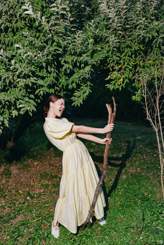 Woman in yellow dress outdoors pulling a large branch with playful energy under trees authenticity inclusivity emphasized candid portrait of joy and movement in nature with sunlight