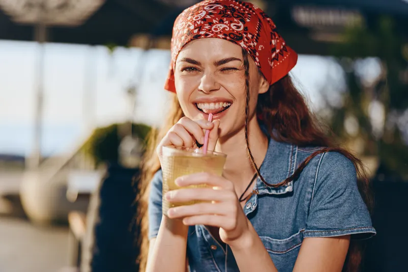 Lifestyle woman in modern boho-western denim street style enjoying a drink outdoors, film color effect enhancing casual and vibrant summer mood.