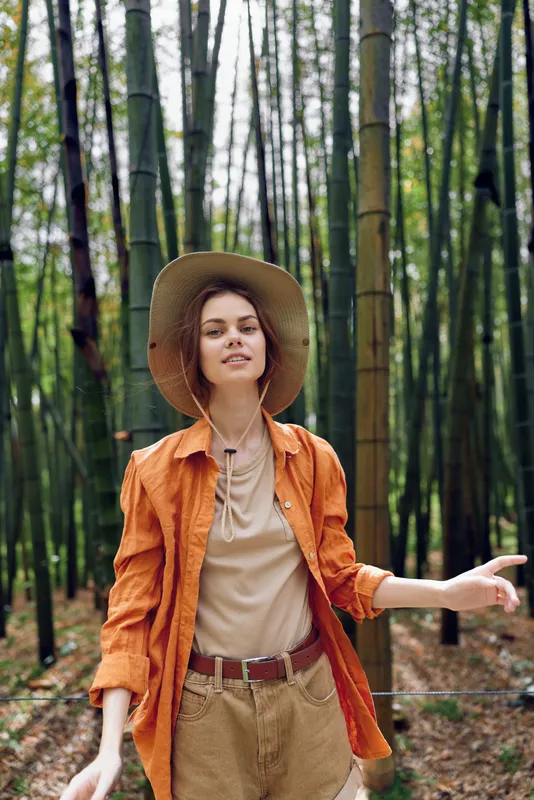 Woman in a bamboo forest wearing a wide hat and casual travel outfit, portrait capturing nature mood and relaxed expression among tall green trees during summer exploration.
