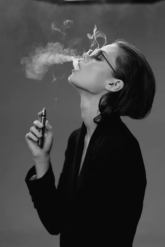 Black and white portrait of a woman vaping with glasses and short hair, exhaling vapor smoke, stylish and modern.