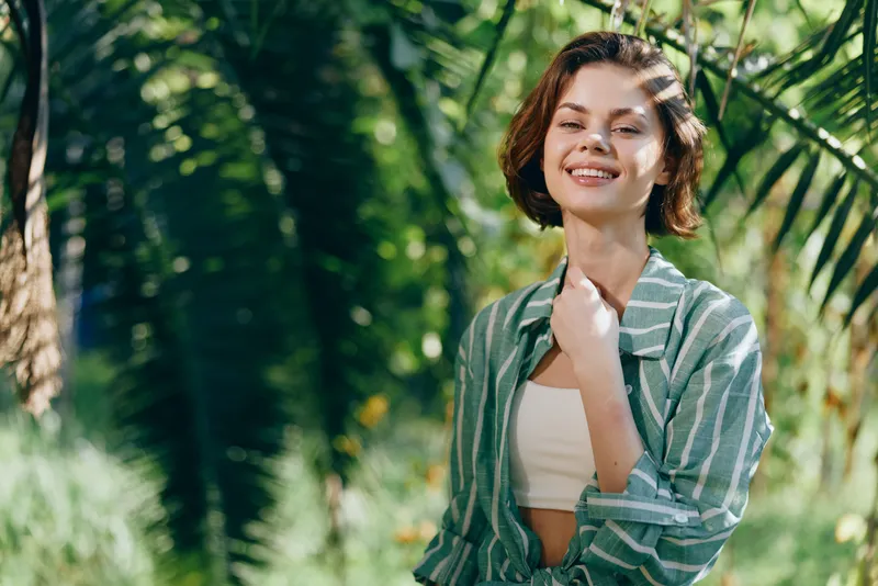 Bright portrait of a cheerful woman outdoors, smiling softly in a striped green shirt under sunny trees.