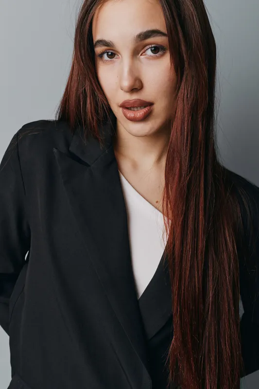 Young woman with long brown hair wearing black blazer and white top posing against neutral background with confident expression and natural makeup for fashion portrait.