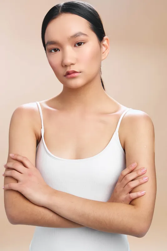 asian woman stands in a soft studio setting wearing a white tank top, arms crossed, a calm confident expression, minimalist background, poised and focused portrait