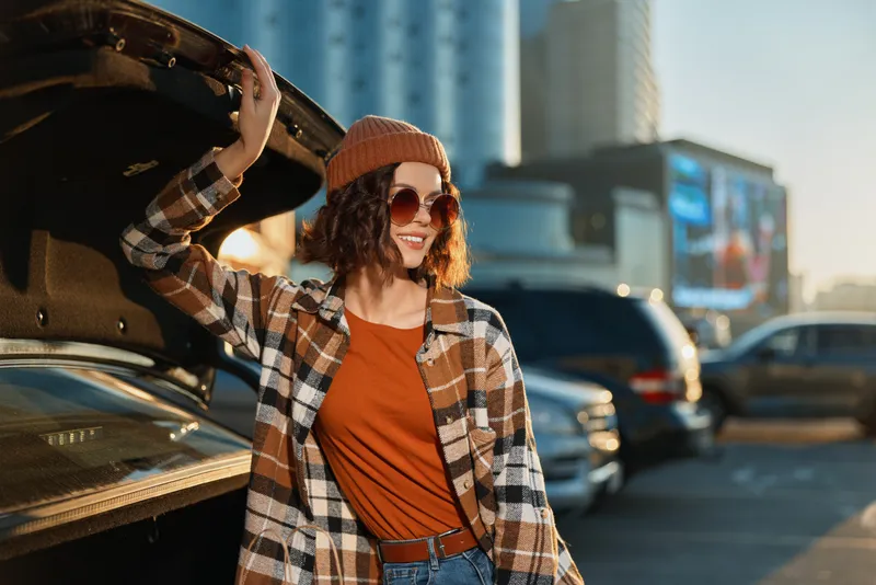 Woman car urban smiling golden hour lifestyle; candid authenticity and mindful living captured at sunset by open trunk, cozy beanie and plaid jacket, emotional storytelling in city