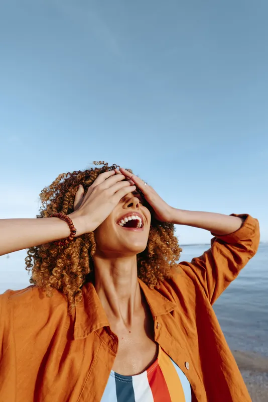 Curly haired woman on a beach laughs with hands over eyes in a sunlit moment