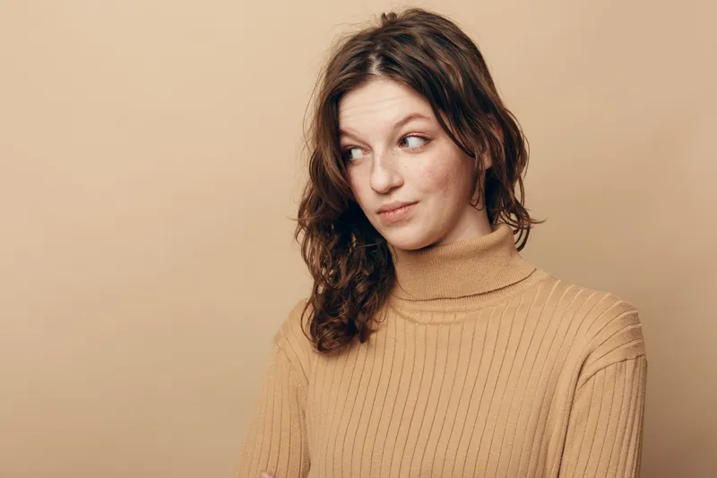 Young woman with curly brown hair wearing beige turtleneck sweater looks sideways with skeptical expression on beige background. Concept of doubt, curiosity, and casual fashion.