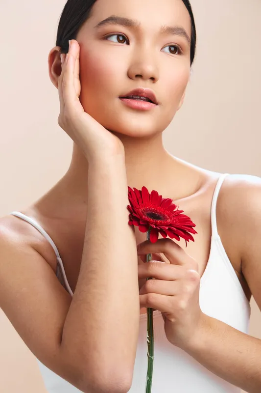 asian woman holds a red flower, delicate pose and thoughtful gaze, a soft portrait conveying grace, beauty, and calm confidence in a studio setting.