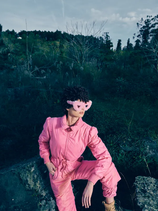 Outdoor portrait of a model in a pink jumpsuit sitting on rocks beside a forest, moody sunlight and calm vibe, fashion focused pose with natural scenery framing