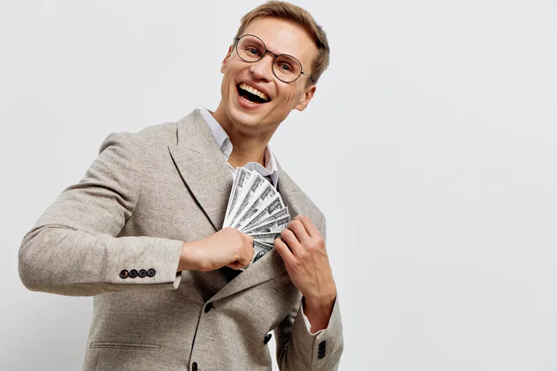 Smiling man dressed in stylish beige blazer holding a fan of cash bills, expressing happiness and success, isolated on plain light background, studio portrait. People lifestyle concept
