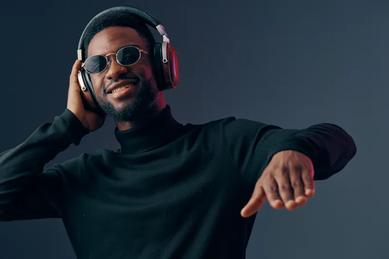 Young Black male with headphones enjoying music, smiling joyfully in a modern black outfit against a dark gradient background, ideal for music and lifestyle concepts