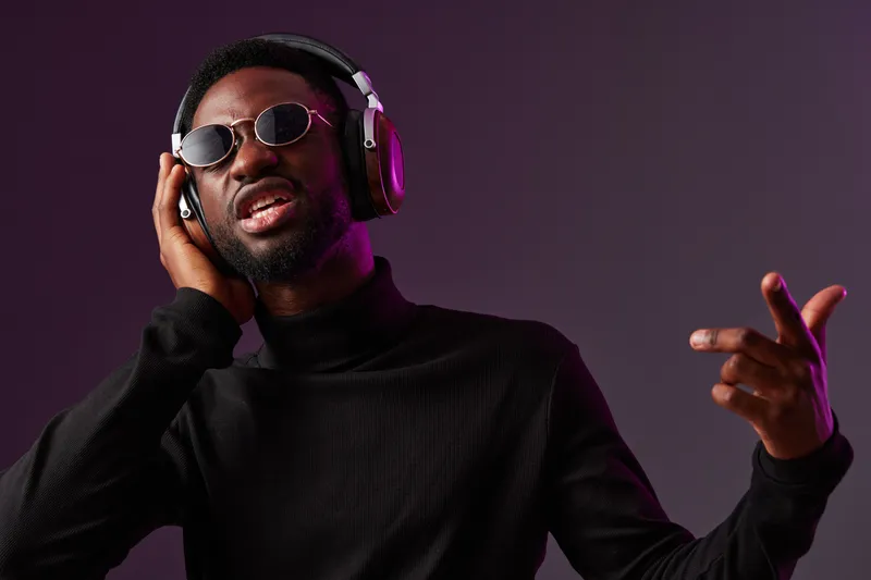 Young Black man enjoying music with headphones, stylish black attire, expressing joy against a dark gradient background