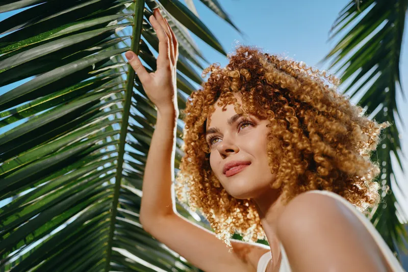 Curly woman enjoying sunny day with tropical palm leaves, blue sky, natural light, and relaxed summer mood in outdoor setting.