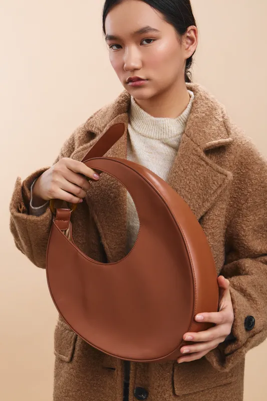 asian woman wearing a beige knit sweater and a brown wool coat holds a round leather handbag, posing in a studio with a calm, stylish vibe and refined fashion sensibility.