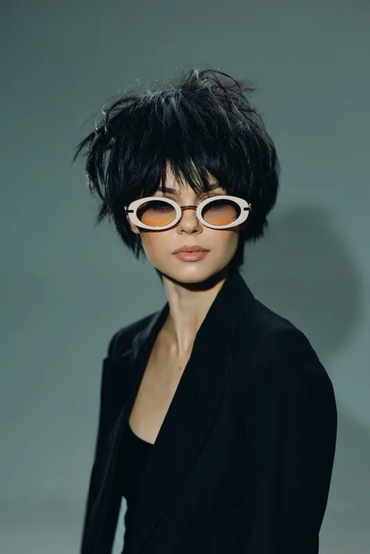 Stylish woman with short black hair and white sunglasses wearing a black blazer, posing confidently against a minimal gray background.