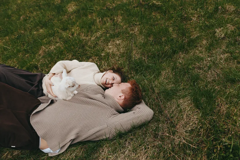 Young couple lies on grass holding a baby goat while sharing a joyful moment outdoors in cozy sweaters during a relaxed lifestyle scene in nature.