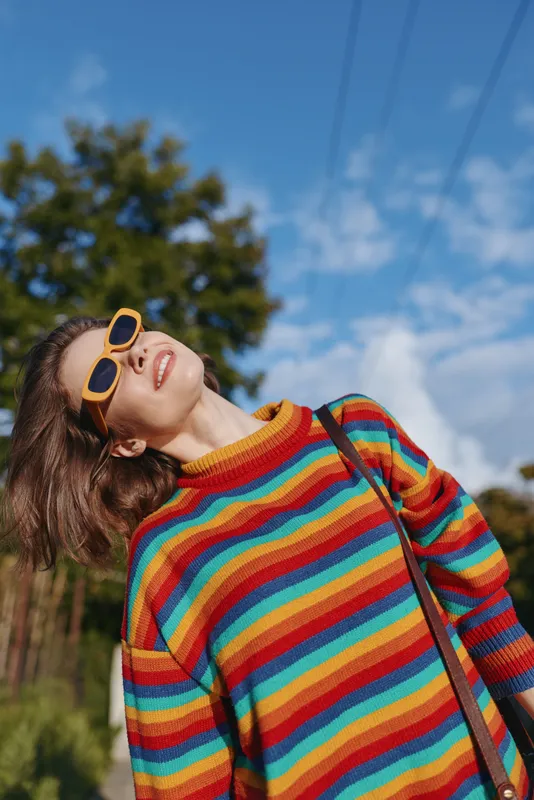 Young woman in a rainbow striped sweater and yellow sunglasses smiles joyfully, leaning back under blue sky in an outdoor park setting; casual outfit, carefree lifestyle and fashion vibe.